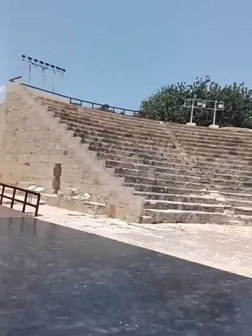 The video shows an ancient amphitheater under a clear blue sky. The amphitheater is constructed of stone seating tiers that curve around a central stage area. Several people are visible in the video. In the upper tiers of the amphitheater, two individuals are seen walking. In the central stage area, a group of people are gathered. One person holds a white umbrella. Another person is bending down, possibly picking something up from the ground. A third person stands a short distance away, looking towards the stage. In the background, trees and stage lighting equipment are visible. The overall impression is of a historical site being visited by tourists on a sunny day.