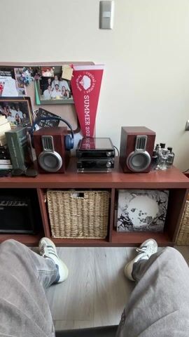 The video shows a person sitting on the floor with their legs outstretched, facing a shelf unit. The shelf unit is against a beige wall. On the top of the shelf unit, there are two wooden-framed speakers, a black stereo system, and a few bottles of liquid. A pair of blue headphones is placed near the left speaker. Behind the shelf, a bulletin board with photos and a pennant that reads "SUMMER 2022" are visible. On the shelves below, there is a small black amplifier, a wicker basket, and a vinyl record. The floor is light gray wood grain. The person is wearing gray corduroy pants and white sneakers with blue stripes. A light switch is visible on the wall above the shelf unit. The video appears to be a static shot with no camera movement.