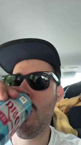 The video shows a man in a baseball cap and sunglasses drinking from a can in the passenger seat of a car. The camera then shows the view from inside the moving car through the front windshield, then a view of the driver. The car is driving down a road lined with trees.