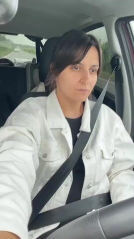 A woman is sitting in the driver's seat of a car, looking forward with a serious expression. She is wearing a white denim jacket over a black top and her seatbelt is fastened. The interior of the car is visible, including the steering wheel and the passenger seat. The background shows a highway with a few cars in the distance, suggesting the car is in motion. The weather appears overcast.