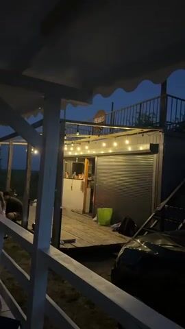 The video shows an outdoor scene at dusk or night. A white railing is in the foreground. Behind it is a wooden deck with a structure on it. The structure appears to be a small building or kiosk with a corrugated metal roll-up door that is partially open. String lights are strung across the front of the structure. A sign with the word "BEREG" is visible above the structure.

On the deck, to the left of the structure, there are some people. One person is sitting and appears to be interacting with something on the ground. To the right of the structure, there is a green bucket and some bags. A portion of a dark-colored car is visible on the right side of the frame. The background shows a dark blue sky with some trees or vegetation. The overall atmosphere is relaxed and casual, possibly indicating a bar, cafe, or gathering spot.