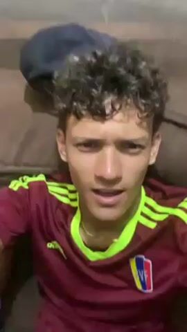 A young man wearing a Venezuela soccer jersey is lying on a brown couch. He starts speaking, touches his face, and then another young man joins him on the couch. The second young man is wearing a red jacket. The camera is close to their faces and the video is shot in a home setting.