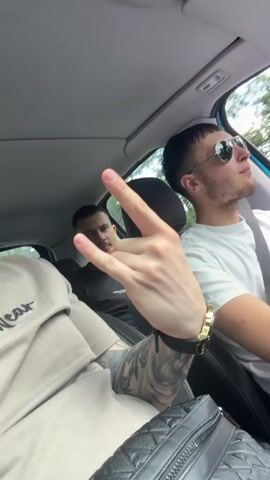 A short video clip captures three young men inside a car during what appears to be daytime. The scene is set from the perspective of a front passenger, looking towards the driver and the back seat.

In the immediate foreground, the left arm of a person is visible, adorned with intricate tattoos. Their hand is raised, making a "rock on" hand gesture. This individual is wearing a light-colored (possibly beige or khaki) t-shirt, with the partial text "Wear" or "W.ear" visible on the fabric. A dark, patterned bag, possibly woven leather, rests on their lap.

The driver, a young man with short hair and a neatly trimmed beard, occupies the mid-ground. He wears a white t-shirt, a gold-toned watch on his left wrist, and mirrored sunglasses, reflecting the car's interior. His gaze is directed towards the right side of the frame, suggesting he is looking out the window or at the road ahead.

In the back seat, a third young man is partially visible, wearing a dark shirt and looking towards the front. The car's interior, including parts of the dashboard and headliner, is visible, along with reflections in the rearview mirror and side mirror. Trees and foliage can be seen through the car windows, indicating movement on a road.

The overall activity involves the group riding in the car. The mood appears relaxed, conveyed by the hand gesture and the driver's calm demeanor. The strong natural light suggests a sunny day. This event is contextualized as taking place in Ruen, Bulgaria.