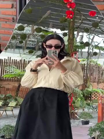 A woman takes a selfie in a mirror. She is wearing a light yellow jacket, a black skirt, and black sunglasses. Her hair is dark and shoulder-length. She is holding a phone in her right hand and using it to take the photo. Her left hand is adjusting her hair. The background of the video includes a brick wall, plants, and outdoor seating. The reflection in the mirror shows the woman and the surrounding environment.