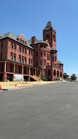 A weathered, red brick building with a prominent clock tower stands under a clear blue sky. The building features a long, covered porch with columns, arched windows, and a multi-story structure with several turrets and dormer windows. In front of the building, a paved road slopes upward. Orange traffic cones and construction materials, including plastic sheeting, are visible near the base of the building, suggesting ongoing repairs or renovations. A small green trash receptacle is also present. The overall scene depicts an old, possibly historical, building with signs of recent activity around its exterior.