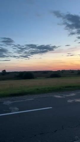 The video displays a time-lapse of a sunset over a rural landscape. The foreground is dominated by a dark asphalt road with a white line marking the lane. In the middle ground, there is a grassy field with scattered bushes and trees. The background features a wide expanse of sky transitioning from blue at the top to vibrant oranges and pinks near the horizon, indicative of a sunset. Wispy clouds are scattered across the sky. The overall impression is one of a peaceful, natural scene captured during the twilight hours.