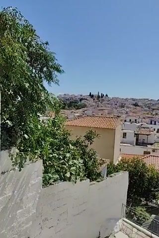 The video shows an aerial view of a town with white buildings and red-tiled roofs under a clear blue sky. In the foreground, there are trees and a white wall. The camera pans to the right, revealing more of the town and a green hillside in the background.