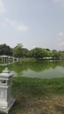 The camera pans across a still lake on a bright, overcast day. On the near shore of the lake, grass can be seen. A small, white decorative stone tower stands on the grass near the edge of the lake. A white, arched bridge spans a part of the lake in the middle-ground. Trees surround the lake, and on the far shore a large, tall stone tower can be seen, reflecting in the lake.