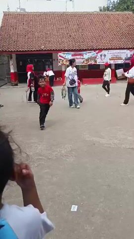 The video shows a group of children and adults in an outdoor courtyard, possibly a schoolyard. The ground is paved with a rough surface. In the background, there is a building with a red-tiled roof and white walls. A banner with red and white stripes, and text that appears to be in Indonesian, is hung across the front of the building. Several people are scattered throughout the courtyard. In the foreground, a young boy wearing a red long-sleeved shirt and black pants walks towards the camera, smiling. Another child in a red shirt and gray pants walks past him from left to right. Further back, an adult in a white shirt and jeans walks, carrying a bag. Another adult, wearing a red headscarf and a white shirt, is seen holding a piece of paper and walking towards the left. Other adults and children are visible in the background, some appearing to be watching or participating in an activity. Some white cones are placed on the ground at intervals. Two pieces of cardboard are lying on the ground near the foreground.