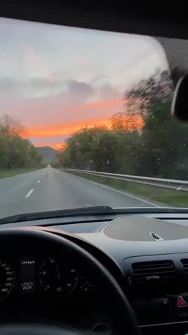 A point-of-view shot from inside a moving car shows the driver's view of a sunset along a rural road. The sky is a mix of orange and pink, framed by trees on either side of the road. The car's dashboard and steering wheel are visible in the foreground. The speedometer reads 125 kilometers per hour. There is music playing in the background.