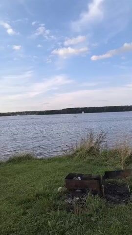 A drone shot pans left over a body of water. The shot is at eye level with the water. There is a forested shoreline in the distance. There are two sailboats on the water. In the foreground, there is grass and a metal barbecue with ash in it. The sky is blue with white clouds.
