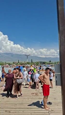 The video shows a wooden pier on a sunny day with a clear blue sky and white clouds. People are walking on the pier and swimming in the turquoise water. Mountains are visible in the distance. In the foreground, a young man in red shorts stands facing the camera. Further down the pier, several women in modest clothing are walking, some carrying bags. Children are also present on the pier and in the water. The scene appears to be a crowded beach or resort area.