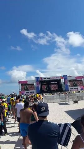 The video shows a crowd of people on a sandy beach under a clear blue sky with scattered white clouds. Many people are wearing brightly colored clothing, predominantly yellow and green, suggesting a sporting event or celebration. In the background, there are large screens displaying images, possibly showing a live broadcast or event information. Some individuals are holding flags or wearing hats. The overall atmosphere appears festive and crowded, with people gathered around the screens or mingling on the beach. The presence of a lifeguard tower-like structure, decorated with what appear to be stars, adds to the beach setting. The camera angle is tilted upwards, emphasizing the vastness of the sky and the number of people below.