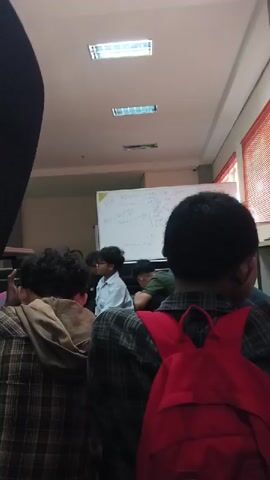 The video shows a classroom setting with several people seated on the floor in front of a whiteboard. The whiteboard has writing on it, but it is not clearly legible. The people appear to be students, and some are looking towards the whiteboard. The room has fluorescent lighting and windows with orange blinds. The camera perspective is from behind the seated individuals, looking forward. The scene suggests a learning or instructional environment.