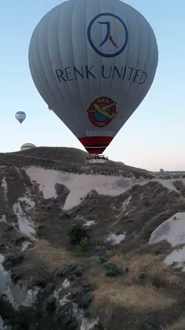 The video captures a scenic aerial view of numerous hot air balloons ascending over a rugged, mountainous landscape. In the foreground, a large white hot air balloon with the inscription "RENK UNITED" and a logo is prominently featured. Below it, a basket with people can be seen. In the middle ground, two more hot air balloons are visible, one yellow and one white with blue accents. The background is filled with a multitude of colorful hot air balloons scattered across the clear blue sky. The terrain below consists of dry, rocky hills with sparse vegetation, suggesting a desert or semi-arid environment. The lighting indicates either early morning or late evening, with soft light illuminating the scene.