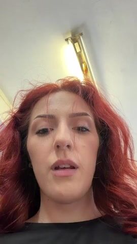 A woman with red hair looks down into the camera. Her hair is slightly messy, and she is wearing a black shirt. She looks directly at the camera with a slight smile. Above her, a fluorescent light fixture is attached to the ceiling. The scene is brightly lit.