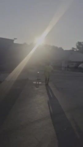 A man in a neon yellow t-shirt and yellow pants walks across a paved area with buildings and trees in the background. The sun is shining brightly, creating a strong lens flare and long shadows. The man is walking away from the camera, and appears to be wearing dark shoes and socks.
