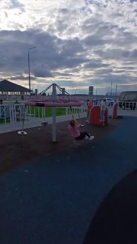 A young girl in a pink shirt and dark pants swings on a playground merry-go-round. Two other children are visible nearby. The sky is cloudy and overcast.