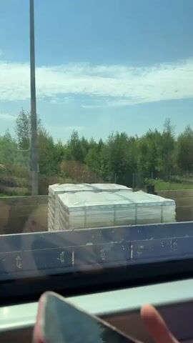 A vehicle is passing a blue truck with a Scania logo, carrying stacks of white items wrapped in plastic. The truck also has metal bars secured in the back. The backdrop features green trees and a partly cloudy blue sky. A phone is partially visible in the foreground.