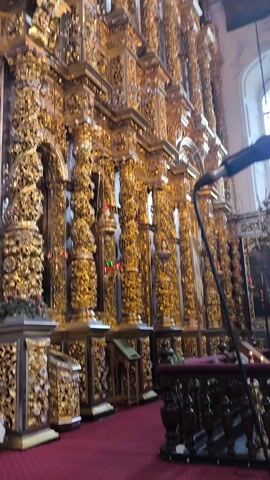 The camera pans up to showcase an ornately decorated wall within a church or cathedral. The wall is covered in gold detailing, featuring intricate carvings and multiple framed images. Light streams in from a window that is visible behind the golden structure. The carvings include depictions of angels and religious symbols. The lighting is relatively dim, highlighting the golden hue of the wall decorations. There are no discernible people in the frame. The video is steady and focuses primarily on the detailed architecture.