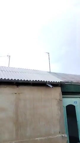 The video shows a view of a cloudy sky above a rooftop. Two antennas are visible on the roof. One antenna is on the left side of the frame, and another is on the right. The roof is made of corrugated material. A wall of a building is visible below the roof. There is a door on the right side of the frame with the number 233 on it.