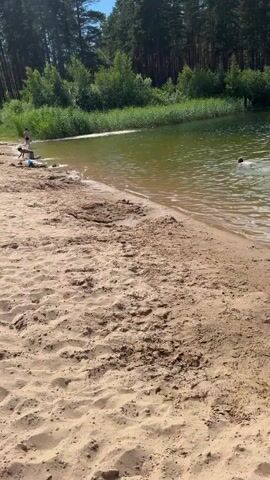 The video shows a sandy beach with a body of water. There are many people spread out on the sand, some are sunbathing, some are playing in the water. There are also beach chairs, umbrellas, towels, children's toys and bicycles on the sand. In the background, there is a forest. The sky is blue with some clouds.