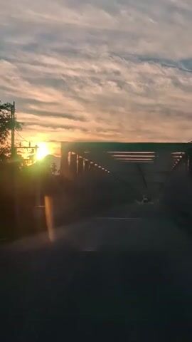 The video shows a view from a vehicle driving across a bridge. The sun is setting on the left side of the frame, casting a warm glow and lens flares. The sky is filled with clouds. The bridge has a dark, metallic structure overhead with a repeating geometric pattern. The road ahead is visible, with a few other vehicles in the distance. The overall mood is serene and atmospheric.