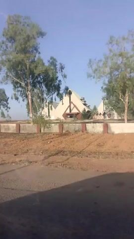 The video depicts a building with a unique architectural design, consisting of multiple pyramid-shaped structures. The building is situated behind a low, concrete wall punctuated with reddish-brown posts. The foreground is characterized by arid, reddish-brown dirt and sparse vegetation. Trees with sparse foliage are scattered around the building, partially obscuring the view. A person walks along a path on the right side of the frame early in the video. The sky is clear and blue. The camera appears to be in motion, likely from inside a moving vehicle.