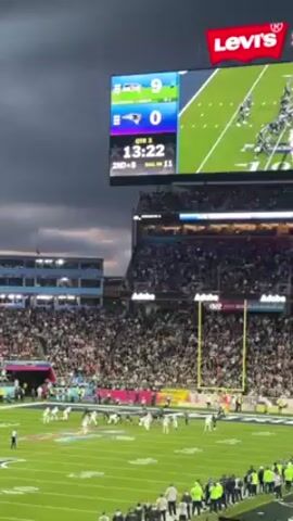 The video shows a football game being played in a stadium. The scoreboard displays the score as Seattle 9 and Patriots 0. The stadium is full of spectators. The game is in the third quarter with 13:18 left on the clock. The team in white is on the field preparing for the next play. The stadium is branded with Levi's and Adobe logos. The sky is cloudy.
