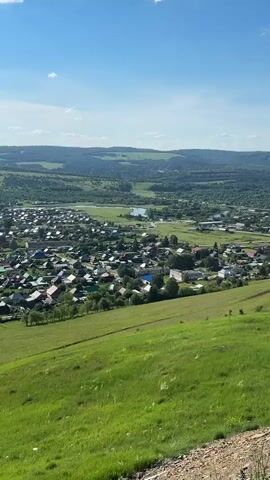 This is a vertical, high-angle, outdoor, long-shot video clip overlooking a rural landscape. The scene is a village nestled in a valley, surrounded by rolling hills covered in lush green trees. In the foreground, a steep, grassy slope dominates, with a small, rocky patch visible at the very bottom. Beyond the slope, the village unfolds with numerous houses featuring colorful roofs, primarily red and blue. Small trees and gardens are scattered throughout the residential area. A body of water, possibly a river or a lake, meanders through the valley behind the village, reflecting the blue sky. Further in the distance, more hills create a layered effect, with the furthest hills appearing hazy and a darker green.

The weather appears to be clear and sunny, with a bright blue sky and a few wispy clouds. The light suggests it's daytime, likely late morning or early afternoon. No people are clearly visible in the clip, and the focus is on the landscape and the village. The overall mood is peaceful and serene, capturing the quiet beauty of a rural summer day. There is no visible text in the clip. The location is identified as Sim, Russia, which aligns with the type of landscape depicted – a Russian countryside with traditional village architecture and natural topography.