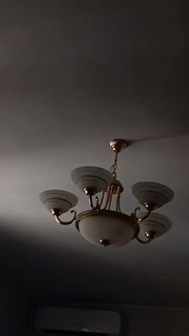 A chandelier with five arms, each holding a frosted glass shade with gold trim, hangs from a white ceiling. The chandelier has a central frosted glass globe and is made of gold-colored metal. Below the chandelier, a white air conditioning unit is mounted on a grey wall. The lighting in the video changes from bright to dark. The video pans slightly to the right, showing a portion of a window with blinds.