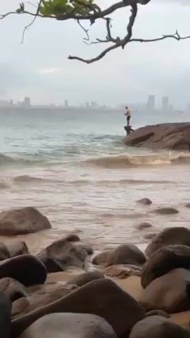 The video depicts a coastal scene with a man fishing from a rock. In the foreground, there are several large, smooth, dark grey rocks scattered across a sandy beach. Gentle waves wash onto the shore, creating a pattern of foamy water against the sand and rocks. In the middle ground, a man stands on a large rock formation, seemingly engaged in fishing. He is silhouetted against the water and the cloudy sky. In the background, a city skyline is visible, with several high-rise buildings punctuating the horizon. The sky is overcast, creating a muted and somewhat grey tone throughout the scene. A tree branch with sparse leaves hangs in the upper left corner of the frame, partially obscuring the view and adding a natural element to the composition.