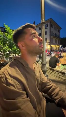 The video shows a young man in a brown shirt sitting outdoors at night. He is looking up and to his right, his expression changing from neutral to a slight smile. In the background, there are people sitting at tables, a building with lights, and trees. The man is looking off-camera, seemingly at something interesting.