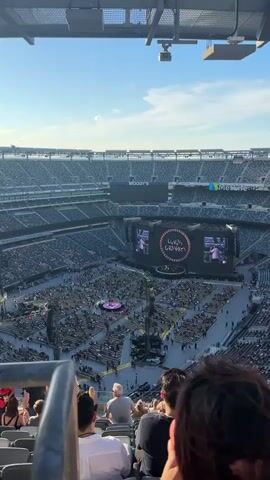 The video shows a wide shot of a large stadium filled with people attending a concert. The stage is set up on the floor of the stadium, with large screens displaying images of performers. The crowd is seated in the stadium's tiered seating, with some people standing. The overall atmosphere appears to be festive and energetic. The time stamp on the video is 00:00 to 00:04.