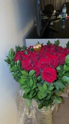 The video shows a hotel room. A bouquet of red roses sits on a desk in the foreground. Behind the desk, a window shows a view of a bridge at night. There are two beds in the room.
