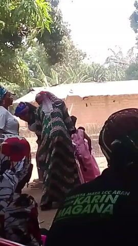 The video shows an outdoor event, likely a wedding or celebration. Several adults are dressed in traditional African clothing, while a group of young girls wear matching pink dresses. The adults appear to be dancing, possibly leading the celebration. The event takes place under the shade of a large tree, with buildings visible in the background. Seated guests are partially visible in the foreground, observing the festivities. The scene suggests a vibrant and culturally rich gathering.