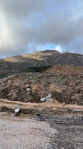 The video shows a construction site with a mountain in the background, under a cloudy sky with a patch of blue. There are piles of dirt and rocks. A red and white striped safety barrier is visible at the left edge. A black pipe lies on the ground. A vehicle then drives into the frame from the left. The ground is muddy.