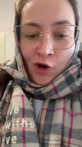 A person wearing a brown puffer coat, a plaid scarf wrapped around their head, and large glasses, holds up a set of keys and points a finger. The background is a light-colored wall and doorway. The person appears to be speaking or singing.