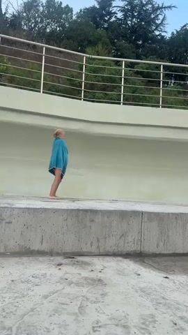 Two young girls are playing by a large concrete structure with a curved, pale green wall. One girl, on the left, is wearing a bright blue towel draped around her shoulders and waist. She is standing on the edge of the concrete structure, looking to the right. The other girl, on the right, is holding a large, colorful cloth, possibly a towel or blanket, which she is waving. She is also on the edge of the concrete structure, facing the first girl. They appear to be interacting playfully. Behind the curved wall, there is a railing, and beyond that, a line of green trees against a pale blue sky. The ground in front of the concrete structure is also concrete and has some puddles of water.