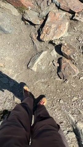 The video shows a first-person perspective of someone standing on a rocky mountaintop. The person is wearing hiking boots and dark pants. The ground is covered in loose rocks and gravel. As the camera pans upward, the view expands to reveal a vast mountain range under a clear blue sky. A winding path can be seen snaking its way down the mountainside. In the distance, a small body of water is visible. The camera tilts upwards further to show a paraglider in the sky.
