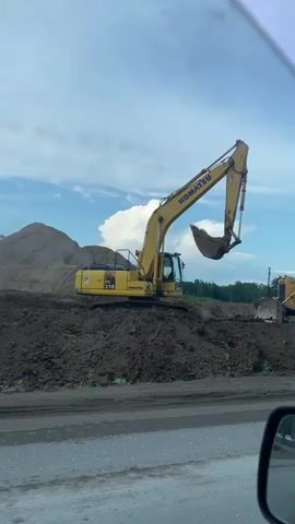 This video shows a construction site with several large pieces of heavy machinery. The main subject is a bright yellow Komatsu excavator, with "KOMATSU" clearly visible on its arm. The excavator is positioned on a mound of dark soil, with its bucket raised high in the air. To the right of the excavator, a yellow bulldozer with the number "5022" visible on its side is also present.

In the background, a large pile of sandy soil is visible, and beyond that, a line of trees. The sky is blue with fluffy white clouds, suggesting it is daytime with fair weather. The scene appears to be an active construction area, with the machinery positioned for earthmoving or excavation.

In the foreground, the reflection of the scene is visible in a car's side mirror. Through the mirror, we can see a road with several cars driving, and in the distance, several tall apartment buildings. This suggests the construction site is located near a populated area, likely in Kabanikha, Russia, as indicated by the location context. No people are directly visible in the video. The overall impression is a typical construction scene on a clear day.