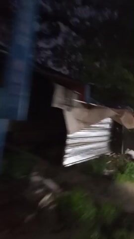 The video shows a blurry view of a street at night. There are several buildings with lit signs on the left side of the street. A motorcycle is visible in the distance, and other vehicles are driving on the road. The ground appears to be wet, suggesting it may have recently rained. The overall atmosphere is dark and somewhat chaotic due to the blurriness of the footage.