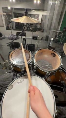 The video shows a person playing a drum set in a recording studio. The drum set is a brown color and has several drums and cymbals. The person is holding a drumstick and hitting the top drum. Other musical instruments, stands, microphones and equipment can be seen behind and around the drum kit. The floor is covered with grey carpet.  A portion of a green wall is seen on the right side. A person can be seen in the background, but their features are not clear. The camera is focused on the drum set, and the person's hand is visible in the foreground.