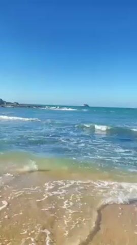 The video opens with a view of a sandy beach and the ocean under a clear blue sky. The water is a mix of turquoise and blue, with gentle waves approaching the shore. The camera pans slightly to the right, and two jet skis appear in the distance. They move towards the camera, each with a rider. The person on the left jet ski wears a light-colored shirt, while the person on the right wears a red shirt. The jet skis are driving through the waves.