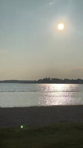 The video shows a serene view of a body of water during what appears to be sunset. The sun, a bright orb, is positioned in the upper right quadrant of the sky, casting a shimmering path of light across the water's surface. The water itself is calm, with gentle ripples. In the foreground, there is a beach composed of small pebbles and a strip of green grass. The horizon is marked by a faint silhouette of trees and buildings on the opposite shore. The sky is a hazy pale blue, with a few wispy clouds. The overall atmosphere is peaceful and tranquil.