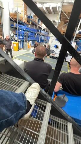 The video shows a warehouse setting. In the foreground, a person is sitting on a metal staircase, wearing jeans and white sneakers. In the background, several people are gathered around a table, appearing to watch a table tennis game. One person is standing with a yellow bin, and another person is sitting in a blue bin. Warehouse shelves filled with items are visible in the background.