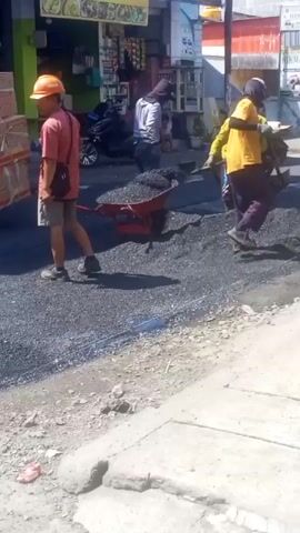 Here is a concise description of the video:

The scene shows a group of construction workers repairing a road on what appears to be a sunny day. One worker in an orange hard hat, a pink shirt, and shorts stands near a red wheelbarrow filled with gravel. Two other workers, one in a yellow shirt and another partially obscured, are actively spreading the gravel on the road surface.  A motorcycle is parked nearby, and shops are visible in the background. The road is freshly covered with the gravel, contrasting with the older concrete sidewalk in the foreground.