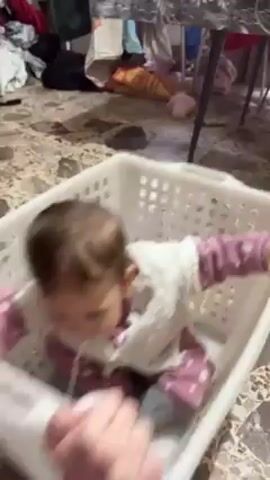 A baby is sitting in a white plastic laundry basket. The baby has brown hair and is wearing pink polka-dot pants, a white shirt, and a white knitted cardigan. A person in a purple shirt is pushing the basket on a marble floor.  The basket is surrounded by clutter, including clothes, bags, and furniture.
