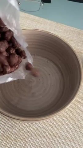 The video shows a close-up, overhead shot of a person pouring chocolate-flavored cereal from a plastic bag into a beige bowl. The cereal is round and appears to be a crunchy variety. The bowl is placed on a textured placemat that is light brown and beige in color. In the background, a pair of glasses is visible, suggesting the person is likely at home or in an office setting. The lighting appears to be natural and soft, indicating it could be daytime. The activity is a simple, everyday one: preparing cereal for a meal. There are no people clearly visible in the frame, only the action of pouring the cereal. The scene is domestic and focuses on the preparation of food. There is no visible text in the video.