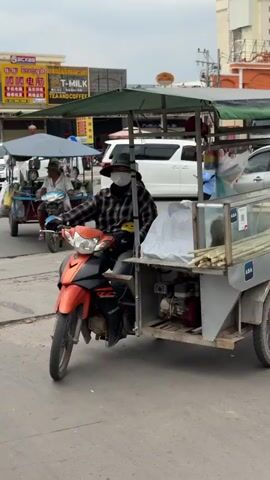 The video shows two men riding motorcycles through a city intersection in Cambodia. The first man drives an orange motorcycle with a sugarcane cart attached, waves to the camera, and turns. The second man rides a blue motorcycle with a cart full of green coconuts. There is another woman riding a red motorcycle. The background shows city streets with storefronts. The sky is cloudy. The street has several holes.